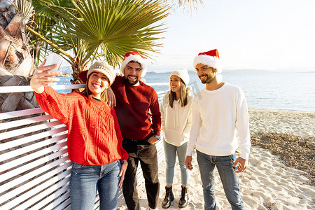 一群戴着圣诞老人帽子的年轻白人朋友在冬天的圣诞节假期在海滩上自拍很开心 — 两对人使用技术分享他们的旅程