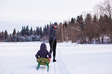 滚动摄影照片_在阳光明媚的冬日里，年轻的爸爸带着他的孩子在雪橇上滚动