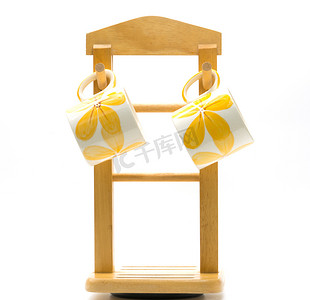 极简主义背景摄影照片_两个咖啡杯外面画着黄色的花，挂在木衣架上，白色背景中带有极简主义风格
