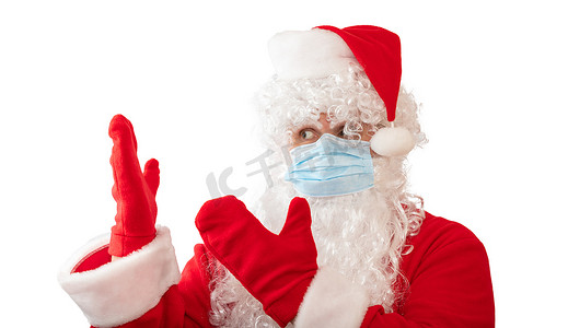 一个穿着圣诞老人服装、戴着医用面具、用双手指着他旁边的东西的男人的视图，在白色背景中被隔离。