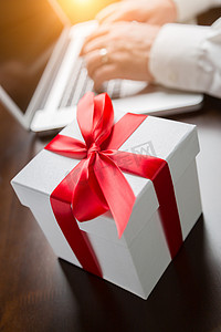 在笔记本电脑上打字的人旁边有红丝带和蝴蝶结的白色礼品盒