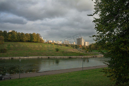河堤公园摄影照片_城市公园的河堤、绿色的草地和大树，天空中灰色的厚云。