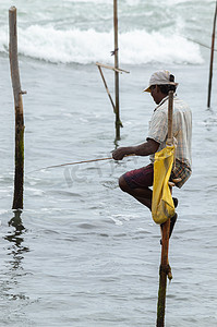 高跷渔夫用他的木杆面向镜头，杆子上有黄色口袋，以斯里兰卡文化中传统独特的方法捕鱼，在海滩上阳光灿烂的夜晚。