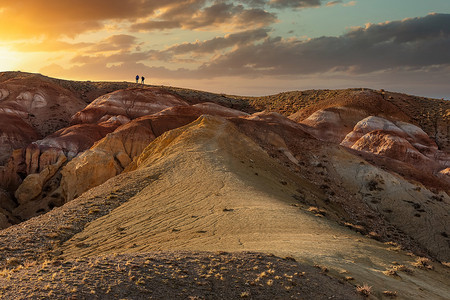 在 Kyzyl-Chin 山谷（也称为火星山谷）巨大的红色山顶上，两个小人物走向日落的美丽低角镜头。