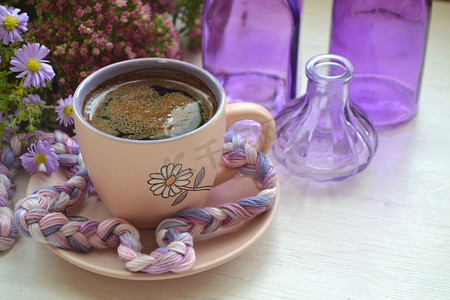 复古杯子里的热咖啡，旧的白色木质背景上，有紫罗兰花和紫色瓶子