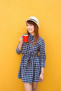 黄色水泥墙背景上穿着裙子、手拿帽子的微笑女人