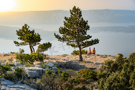 背大石头摄影照片_一对成熟的夫妇坐在一块大石头上，在日落时俯瞰海滨，望着地平线