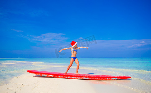 度假时戴着圣诞帽的可爱小女孩在冲浪板上练习冲浪姿势