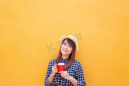 微笑的女人穿着裙子，戴着帽子，在黄色水泥墙背景上拿着红色咖啡杯，思考，想象