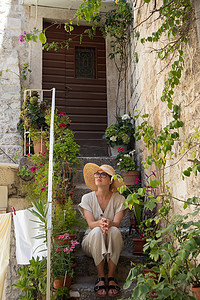 手机夏季夏季摄影照片_美丽的女游客戴着大草帽太阳帽和太阳裙坐在旧石屋楼梯上放松在炎热的夏日地中海成本的夏季旅行中。