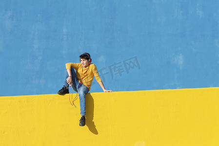 一个穿着休闲服的小男孩坐在黄色围栏上靠着蓝色墙壁，同时用手机通过耳机听音乐的正面图