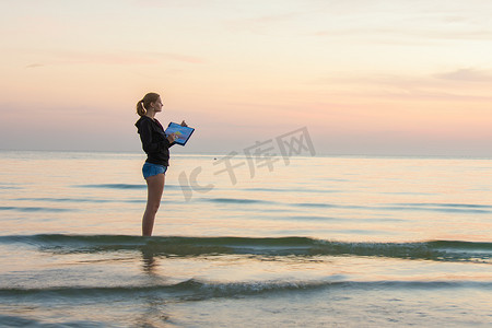 小女孩站在沙滩上的水中，在一张纸上画上夕阳的颜色