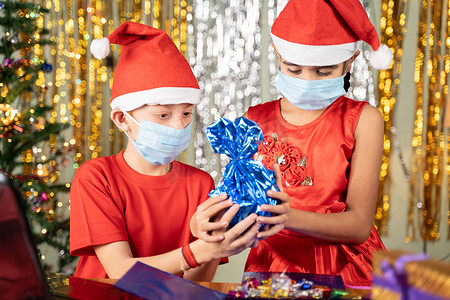戴着医用面具的孩子们在笔记本电脑前在圣诞前夕在家里打开带有装饰背景的礼物 — 由于冠状病毒或 covid-19 大流行而导致遥远的圣诞节庆祝活动的概念。