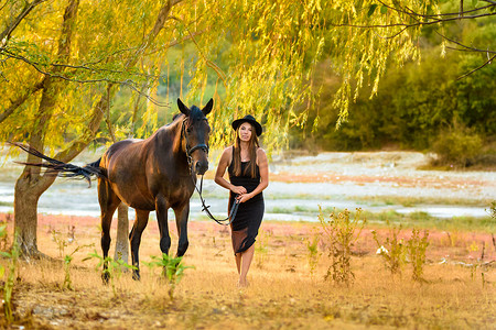 带帽子的女孩摄影照片_一个穿着漂亮的黑色连衣裙和一顶黑色帽子的女孩带着一匹马穿过田野