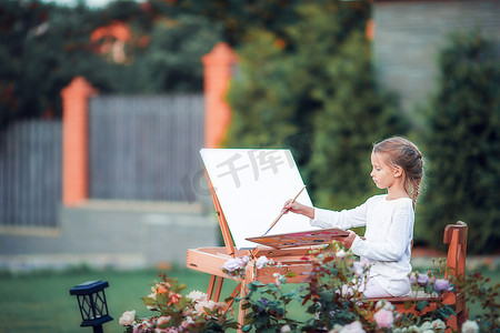 温暖创意画摄影照片_可爱的小女孩在户外温暖的日子里在画架上画画