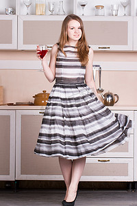 女人酒摄影照片_年轻漂亮的女人穿着裙子，在她的厨房里拿着一杯酒。