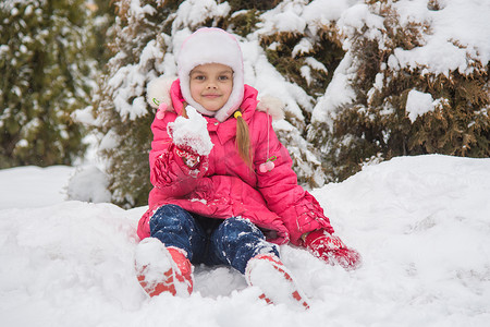 七岁的女孩坐在雪地里，雪让他手里拿着雪