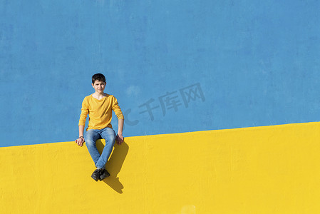 蓝色的牛仔裤摄影照片_阳光明媚的日子里，一个穿着休闲服的小男孩坐在黄色的篱笆上，靠着蓝色的墙壁