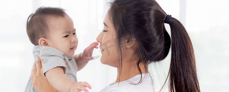 年轻的亚洲母亲在家里的卧室里抱着小女婴，妈妈带着孩子和照顾孩子，女人和孩子无忧无虑，蹒跚学步的孩子和父母，情感和表达，家庭观念。