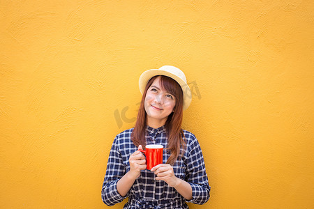 微笑的女人穿着裙子，戴着帽子，在黄色水泥墙背景上拿着红色咖啡杯，思考，想象