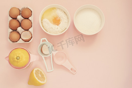 用于烹饪柠檬蛋糕或糖果的配料和厨房烘焙工具 — 鸡蛋、面粉、糖、手榨汁机，背景为柔和的粉红色。