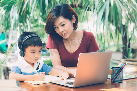 年轻的亚洲母子使用笔记本电脑在家学习和学习，男孩在笔记本上写作业并戴耳机，老师或妈妈支持孩子，教育理念。