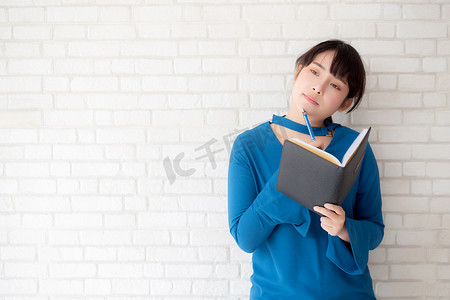 美丽的亚洲女人微笑着站在家里的混凝土水泥白色背景上思考和写笔记本，女孩在书本、教育和生活方式概念上的家庭作业。