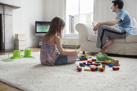 女孩坐在地板上玩积木，父亲在家看电视