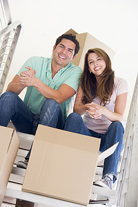 楼梯上搬箱子摄影照片_一对夫妇坐在楼梯上，带着箱子在新家微笑