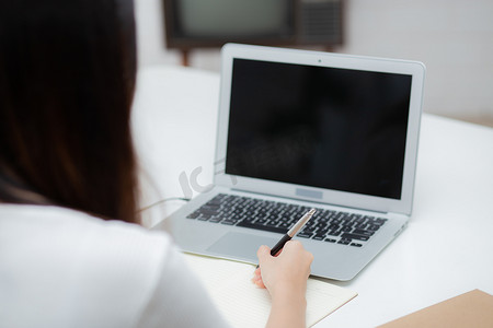 学习规划摄影照片_年轻的亚洲女性在笔记本上写作以规划工作和在家里的办公桌上使用笔记本电脑空白屏幕、女孩笔记、女性学习和学习、商业和沟通概念。