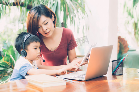 年轻的亚洲母子使用笔记本电脑和平板电脑在家学习和学习，男孩戴着耳机进行远程电子学习，老师或妈妈支持孩子，教育理念。