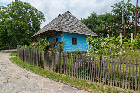 一座美丽的房子，墙壁是用木头做成的彩绘墙，站在木板栅栏后面，院子里种着绿色植物，旁边是一条石头铺成的街道。