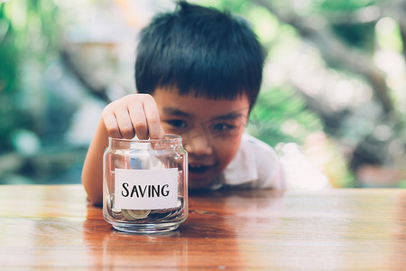可爱数钱摄影照片_快乐的亚洲儿童存钱把硬币放在玻璃杯中，以获得未来的财富和收入增长、金融和投资以取得成功、男孩计划和存款、商业和经济概念。