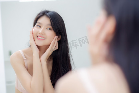 奶油摄影照片_美丽的亚洲年轻女性微笑着看着镜子，用护肤品和化妆品检查脸部，以恢复活力和卫生，美丽的女孩用奶油或乳液清洁面部，并保持健康。