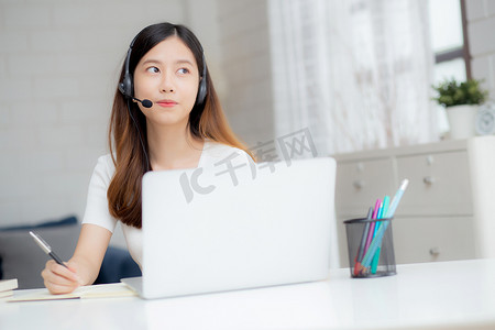 年轻的亚洲女性戴着耳机在线学习，在办公桌上进行电子学习，戴着耳机的女孩在家里用笔记本电脑学习互联网，新常态，远程教育和培训。