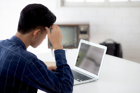 年轻的亚洲商人在笔记本电脑上工作时头痛，显示空白屏幕，最后期限在家，失败的商人筋疲力尽，压力大，担心，沮丧和失败。