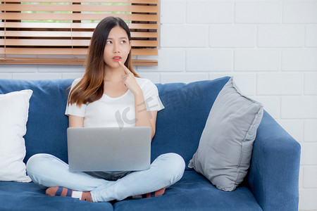 美丽的年轻亚洲女性在家里的沙发上使用笔记本电脑工作并思考想法，自由职业者女孩坐在沙发上使用笔记本电脑在客厅上网，一个人，生活方式概念。