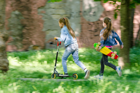 一个女孩摄影照片_女孩骑着滑板车开心地走着，后面跟着另一个女孩，手里拿着溜冰鞋