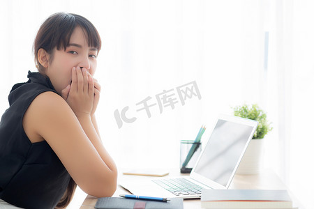 美丽的年轻自由亚洲女性在办公室的笔记本电脑上工作无聊和疲倦，女孩懒惰困倦，在笔记本上打哈欠，女秘书疲惫不堪，商业理念。