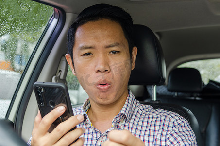 身穿格子衬衫的亚洲男子在车里看着智能手机。