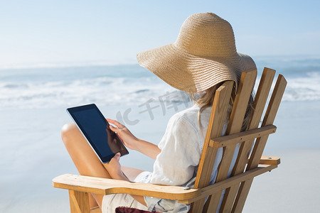 微笑的金发女郎坐在海边的木制躺椅上使用平板电脑