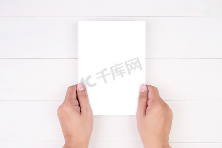 a4模板摄影照片_年轻人的手拿着海报或卡片或小册子，木桌上有白纸空白、贺卡、a5、a4、传单广告和设计空、复制空间、模型模板的消息。