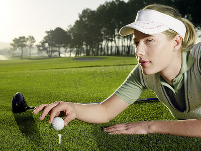 多人高尔夫摄影照片_美丽的年轻女高尔夫球手躺在草地上，同时将球放在发球台上