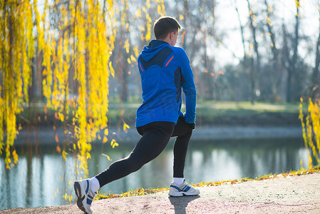 年轻的男跑步者在寒冷的阳光明媚的秋天早晨在公园里伸展。