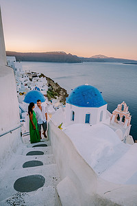 希腊圣托里尼岛摄影照片_希腊圣托里尼，年轻夫妇在圣托里尼岛享受豪华假期，在日出期间观看蓝色圆顶教堂和粉刷成白色的希腊伊亚圣托里尼村的日出，男人和女人在希腊度假