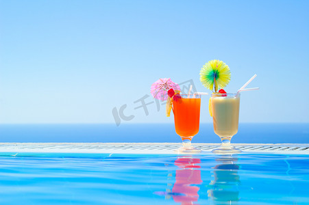 蓝色大海摄影照片_在温暖的蓝色大海背景下的游泳池附近的两个五颜六色的热带鸡尾酒。
