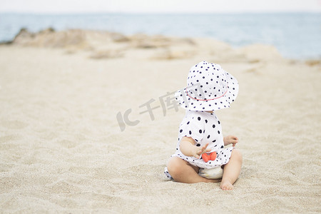 可爱的小女孩，穿着裙子，戴着波尔卡圆点上的帽子，坐在海边的沙滩上，望着大海