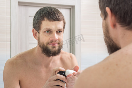 一个胡子拉碴的男人在手掌上涂上剃须泡沫，然后照着浴室里的镜子