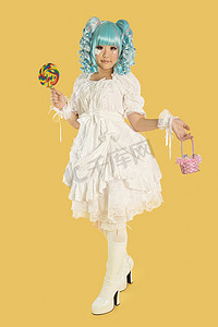 手持棒棒糖摄影照片_黄色背景下打扮成洋娃娃的年轻女子的全长肖像，手持棒棒糖
