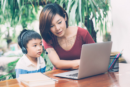 年轻的亚洲母子使用笔记本电脑在家学习和学习，男孩戴着耳机进行远程电子学习，老师或妈妈支持孩子，教育理念。